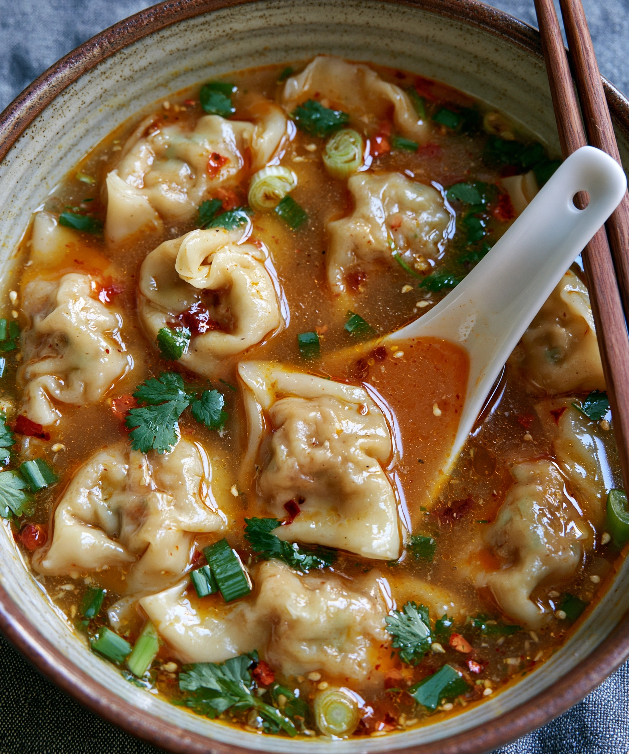 Easy dumpling soup recipe with plump wontons in a savory ginger broth with chili oil, sliced green onions, cilantro and sesame seeds, served with chopsticks and a soup spoon