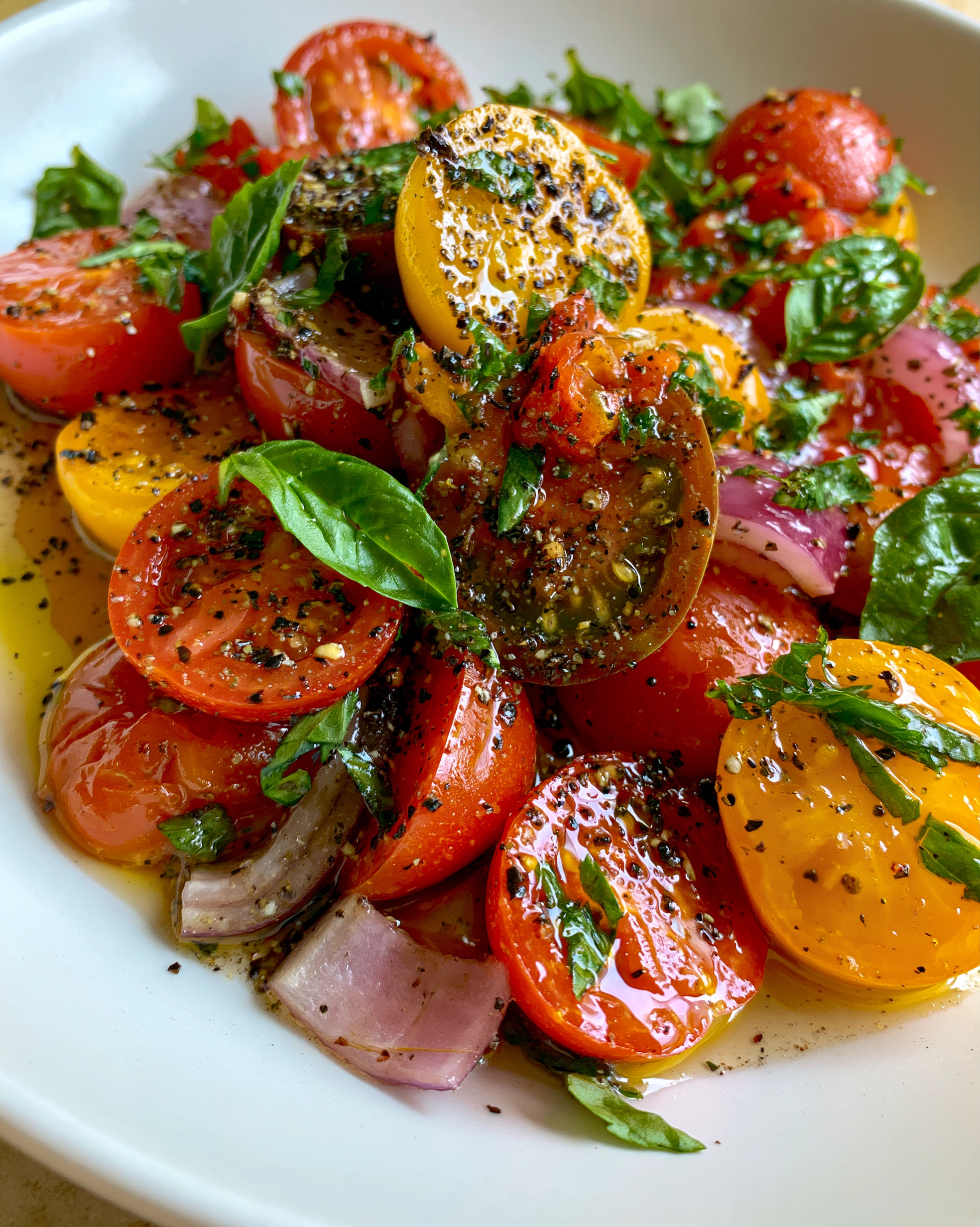Easy tomato salad in a white bowl with halved red, yellow, and dark cherry tomatoes, sliced red onion, fresh basil, and cracked black pepper in a light olive oil dressing