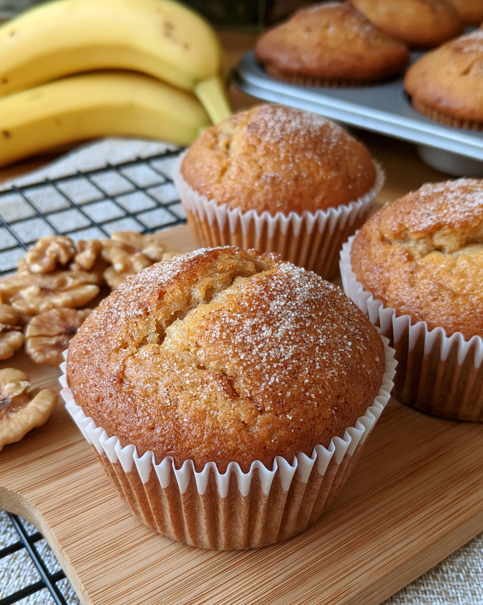 Easy healthy banana muffins recipe with tall golden domes and a light sugar sparkle, bananas and walnuts beside them