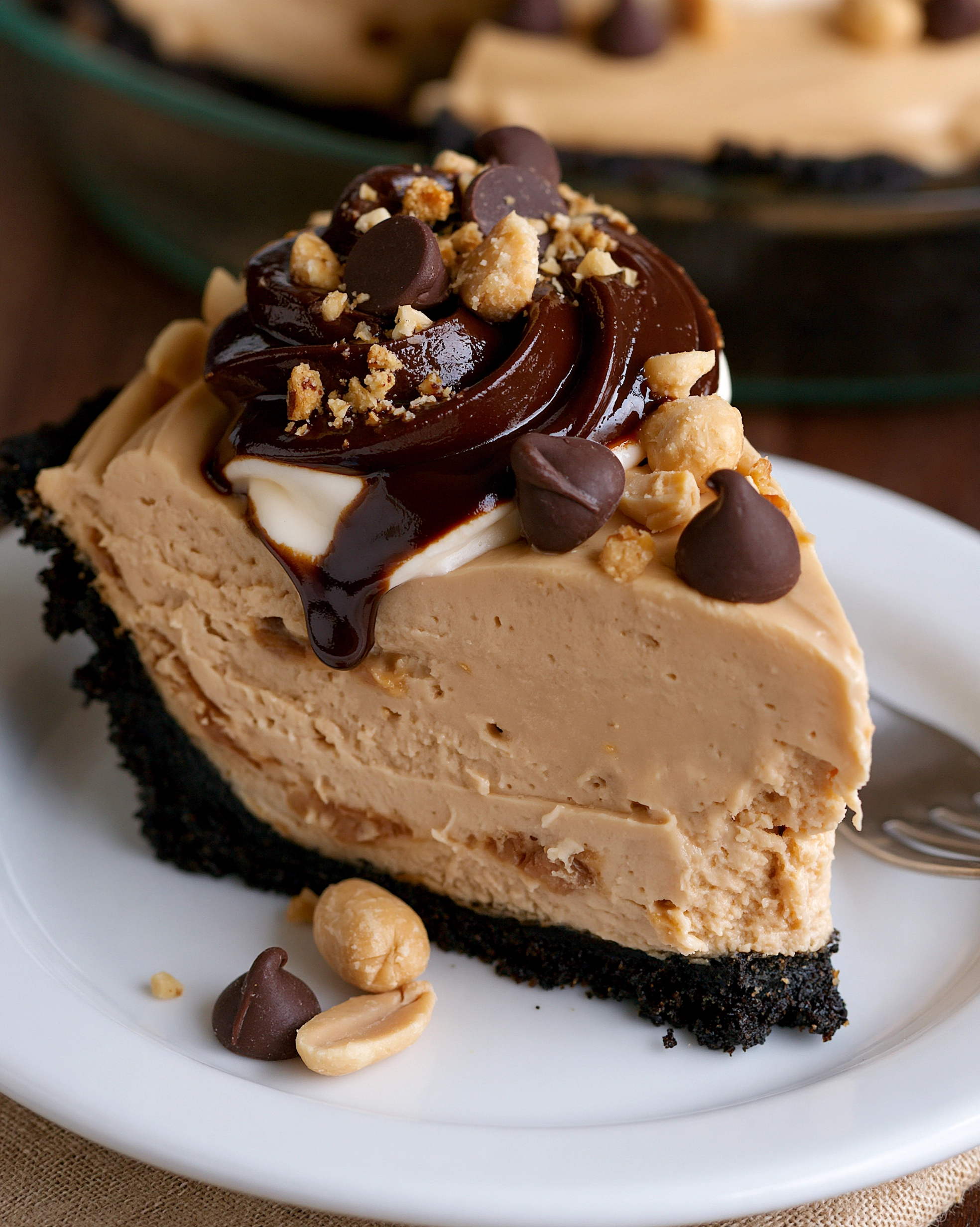Easy no-bake peanut butter pie slice with a dark Oreo crust, creamy peanut butter mousse, glossy chocolate ganache swirls, chocolate chips and chopped peanuts